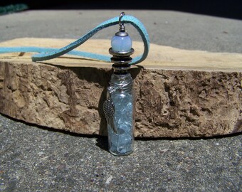 May include: A silver pendant necklace with a glass vial filled with blue crystals. The vial is topped with a silver cap and a light blue opal bead. The necklace is on a light blue suede cord.