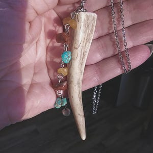 May include: A necklace with a long, light brown antler pendant and a silver chain. The pendant is adorned with a string of small, colorful stones, including turquoise and orange.