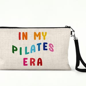 Puede incluir: Una bolsa de maquillaje de lona beige con una cremallera negra y una correa negra. La bolsa tiene el texto "IN MY PILATES ERA" impreso en colores arco&iacute;ris.