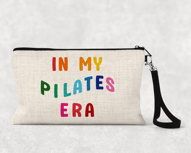 Puede incluir: Una bolsa de maquillaje de lona blanca con una cremallera negra y una correa negra. La bolsa tiene el texto "In My Pilates Era" impreso en colores arco&iacute;ris.