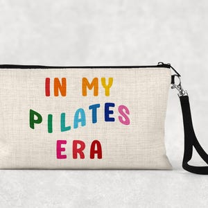 Puede incluir: Una bolsa de maquillaje de lona blanca con una cremallera negra y una correa negra. La bolsa tiene el texto "In My Pilates Era" impreso en colores arco&iacute;ris.