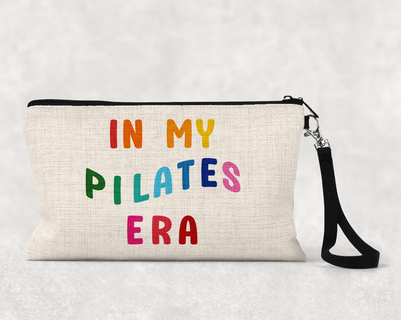 Puede incluir: Una bolsa de maquillaje de lona blanca con una cremallera negra y una correa negra. La bolsa tiene el texto "In My Pilates Era" impreso en colores arco&iacute;ris.