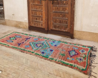 May include: A colorful, hand-woven rug with a geometric pattern in shades of red, green, blue, and purple. The rug is laid out on a tiled floor in front of a wooden door.