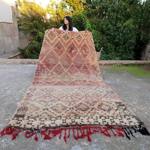 May include: A large, handwoven rug with a geometric pattern in shades of red, brown, and beige. The rug has a fringe border and is being held up by a person.