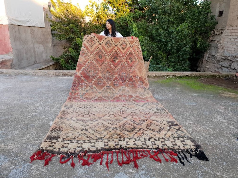 May include: A large, handwoven rug with a geometric pattern in shades of red, brown, and beige. The rug has a fringe border and is being held up by a person.