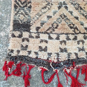 May include: A beige and brown Moroccan rug with a geometric pattern. The rug has red fringe along the edges.