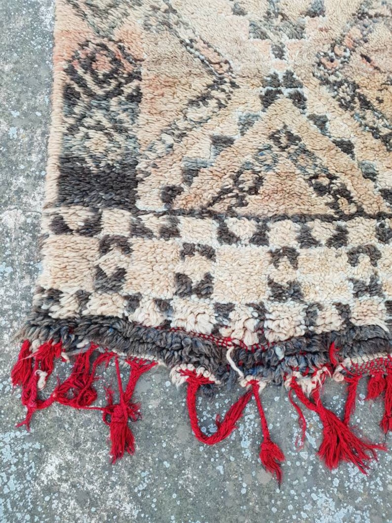 May include: A beige and brown Moroccan rug with a geometric pattern. The rug has red fringe along the edges.