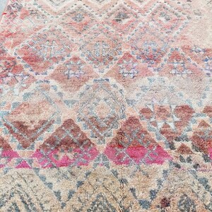 May include: A vintage Moroccan rug with a geometric pattern in shades of pink, brown, and gray. The rug has a fringe border.