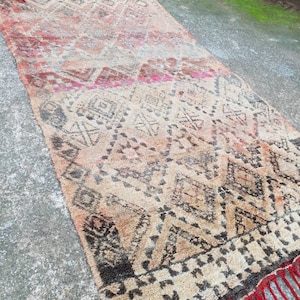 May include: A vintage Moroccan rug with a geometric pattern in shades of brown, beige, and red. The rug has a fringe border and is made of wool.