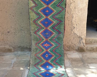 May include: A long, green rug with a diamond pattern in shades of blue, orange, and white. The rug has a fringe on the bottom.