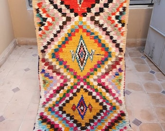 May include: A colorful Moroccan rug with a geometric pattern in shades of red, yellow, blue, green, and pink. The rug has a fringe border and is woven with a thick, fluffy pile.