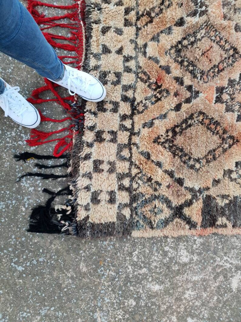 May include: A beige and brown rug with a geometric pattern and red and black fringe. The rug is on a concrete floor.