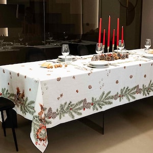 May include: A rectangular dining table covered with a white tablecloth featuring a printed pattern of green pine branches, brown pinecones, and small squirrels. The table is set with white plates, clear glasses, and a central row of tall red candles.