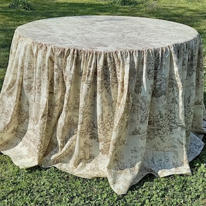 Può includere: Un tavolo rotondo coperto da una tovaglia color crema con un disegno di scena pastorale marrone. La tovaglia scende fino a terra, creando un effetto a gonna ampia. Il tavolo è posto su un prato verde.