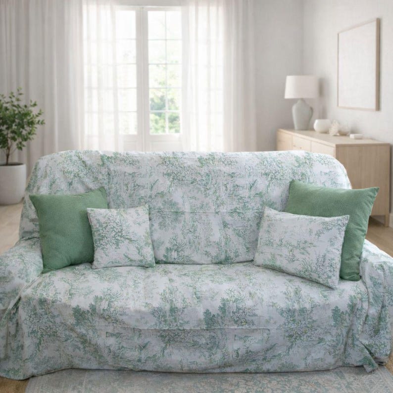 Puede incluir: Un sof&aacute; cubierto con una tela blanca que presenta un patr&oacute;n toile verde de &aacute;rboles y paisajes. El sof&aacute; est&aacute; decorado con dos cojines cuadrados de color verde s&oacute;lido y dos cojines rectangulares que combinan con la tela toile verde y blanca.