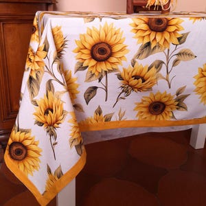 May include: A white tablecloth featuring a repeating pattern of yellow sunflowers with brown centers and green leaves. The fabric has a solid yellow border along the bottom edge, showcasing a floral design printed across the entire surface.
