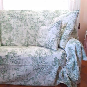 Puede incluir: Un sof&aacute; cubierto con una tela blanca que presenta un patr&oacute;n repetitivo de toile de jouy en verde salvia, con escenas pastorales de &aacute;rboles, follaje y figuras. El conjunto incluye un coj&iacute;n cuadrado a juego contra el respaldo.