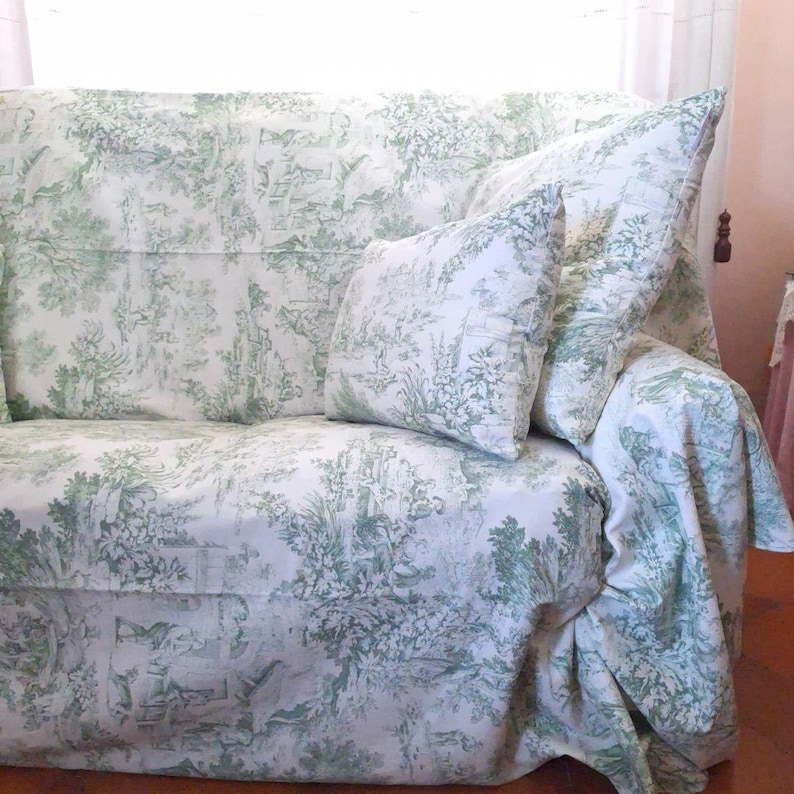Puede incluir: Un sof&aacute; cubierto con una tela blanca que presenta un patr&oacute;n repetitivo de toile de jouy en verde salvia, con escenas pastorales de &aacute;rboles, follaje y figuras. El conjunto incluye un coj&iacute;n cuadrado a juego contra el respaldo.