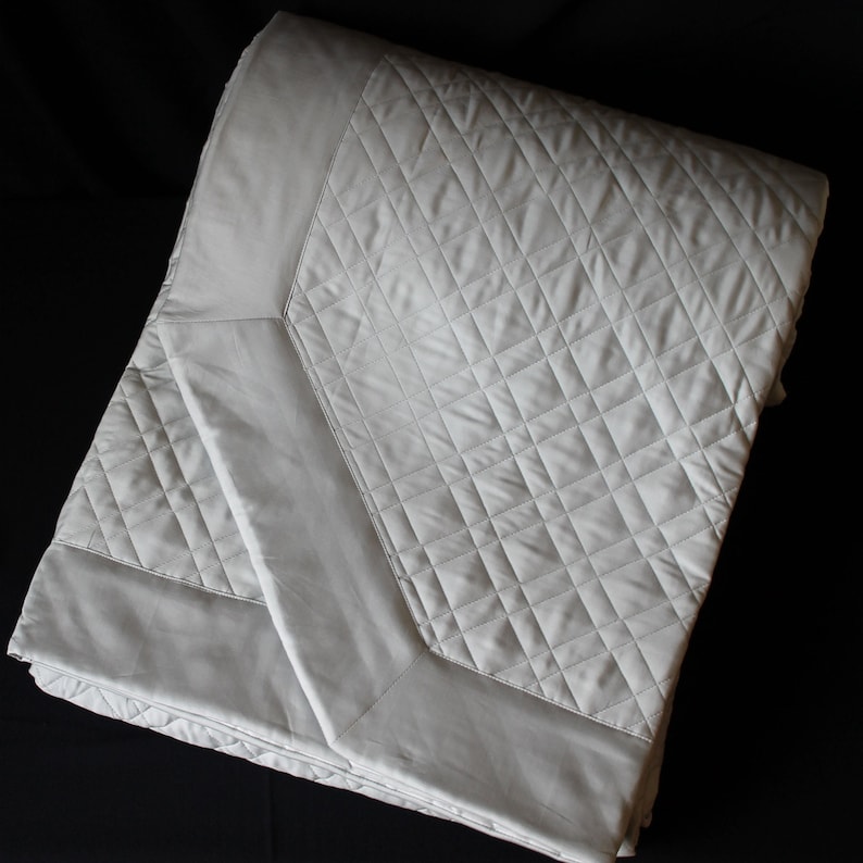 May include: A light gray quilted bedspread featuring a diamond-stitched pattern across the center panel, bordered by a smooth, non-quilted fabric frame. The textile has a subtle sheen and is folded to display both the quilted and flat sections.