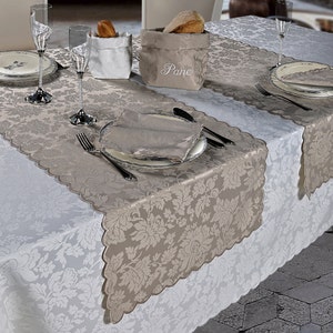 Puede incluir: Un camino de mesa beige con borde festoneado y estampado floral adamascado, colocado sobre un mantel blanco. Una panera de tela beige con la palabra Pane bordada en blanco se encuentra sobre la mesa junto a platos, vasos y cubiertos.