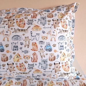 May include: White pillowcase featuring a repeating pattern of various cartoon cats in different poses and colors. The cats are depicted with playful expressions and are interspersed with paw prints, fish bones, and yarn balls. The word "cat" is printed on a cat food bowl.