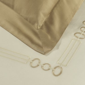 May include: A beige pillowcase with a smooth finish and a wide border, featuring an embroidered geometric pattern of interlocking circles and rectangles in a lighter cream thread along the edge of the fabric on a plain off-white background.