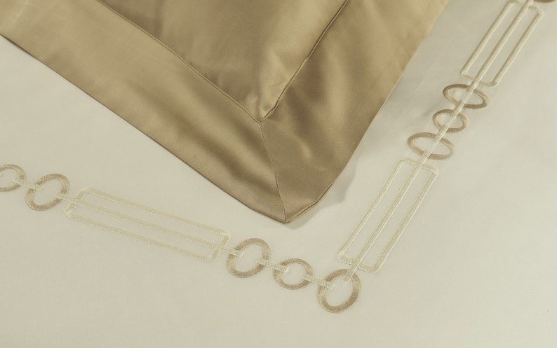 May include: A beige pillowcase with a smooth finish and a wide border, featuring an embroidered geometric pattern of interlocking circles and rectangles in a lighter cream thread along the edge of the fabric on a plain off-white background.