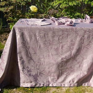 May include: A light purple tablecloth featuring a tonal damask floral pattern with a subtle sheen. Several matching folded napkins are arranged on the surface of the fabric, which drapes over the edges of a rectangular table in an outdoor setting.
