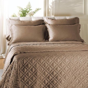 May include: A solid taupe bed set featuring a quilted coverlet with a diamond-stitched pattern and four matching rectangular pillow shams. The bedding has a smooth, slightly lustrous finish and is arranged neatly on a bed frame with a headboard.