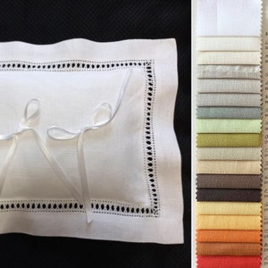 May include: A rectangular white linen pillow featuring a decorative hemstitch border and two thin white ribbon ties. Beside the pillow is a vertical color swatch card displaying a variety of fabric samples in shades of white, gray, green, brown, yellow, orange, and red.