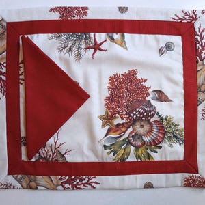 Peut inclure: Un set de table rectangulaire à fond crème orné d'un motif de corail rouge et de coquillages, encadré par une bordure rouge unie. Une serviette triangulaire rouge assortie est pliée et posée sur le côté gauche du set de table.
