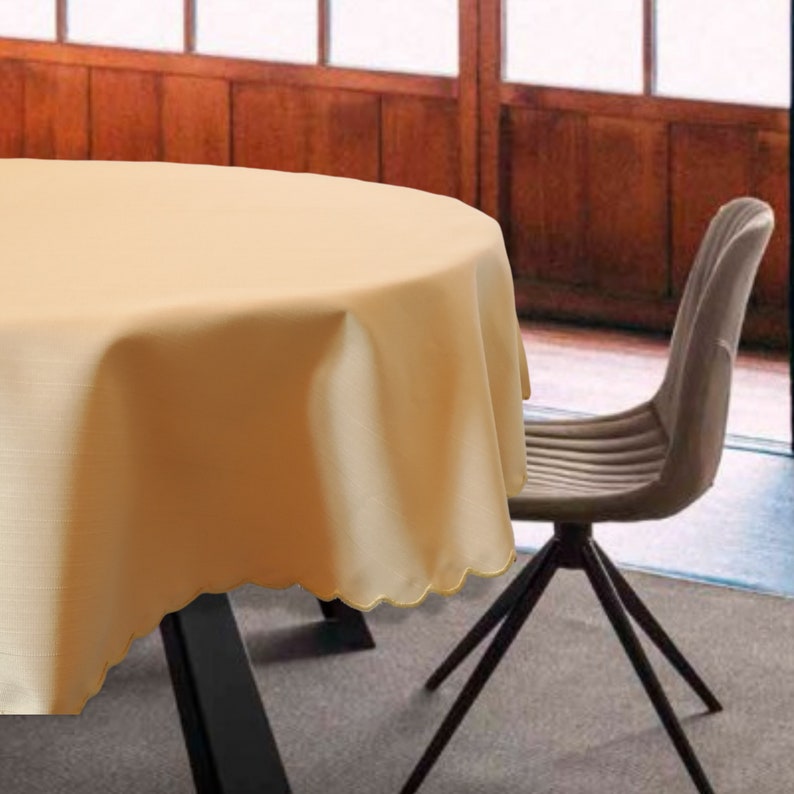 May include: A round, solid light beige tablecloth with a scalloped edge draped over a table. The fabric features a subtle, fine-textured weave pattern throughout, providing a clean and uniform appearance across the entire surface of the round cover.