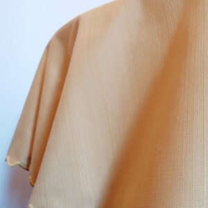 May include: A light tan or beige fabric with a subtle vertical striped texture and a scalloped edge trim. The material appears smooth with a slight sheen, featuring a consistent weave pattern throughout the surface and a finished border detail.