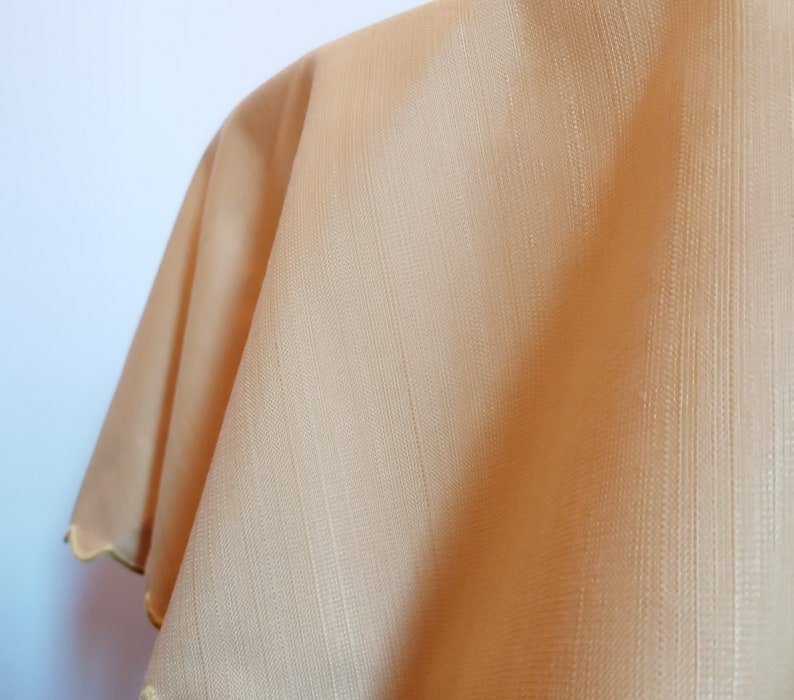 May include: A light tan or beige fabric with a subtle vertical striped texture and a scalloped edge trim. The material appears smooth with a slight sheen, featuring a consistent weave pattern throughout the surface and a finished border detail.