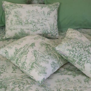 May include: Three rectangular and square pillows featuring a white fabric base with a repeating green toile de jouy pattern depicting pastoral scenes with figures, trees, and buildings. The pillows are arranged on a matching green patterned fabric.
