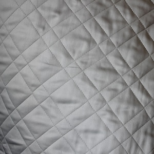 May include: A solid light gray fabric featuring a repeating diamond-shaped quilted pattern. The surface is composed of stitched lines that intersect to form uniform geometric shapes across the entire visible area of the textile material.