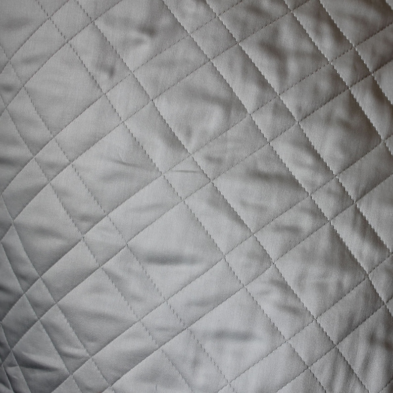 May include: A solid light gray fabric featuring a repeating diamond-shaped quilted pattern. The surface is composed of stitched lines that intersect to form uniform geometric shapes across the entire visible area of the textile material.