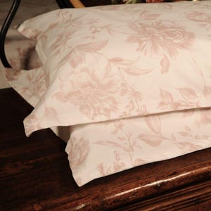May include: Two white pillowcases with a large, repeating floral pattern in a soft rose color. The design features detailed botanical illustrations of flowers and leaves, creating a romantic and elegant aesthetic. The pillowcases are neatly stacked.