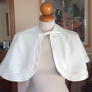 May include: A solid white satin capelet featuring a rounded neckline and a delicate ribbon tie closure at the front. The garment has a smooth, lustrous finish with a wide, stitched border detail along the curved hemline, draping over the shoulders.