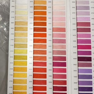 May include: An Aurifil thread color chart featuring a grid of numbered swatches in shades of yellow, orange, pink, red, and purple. The white card displays columns of thread samples arranged by color family, each labeled with a specific numerical code.