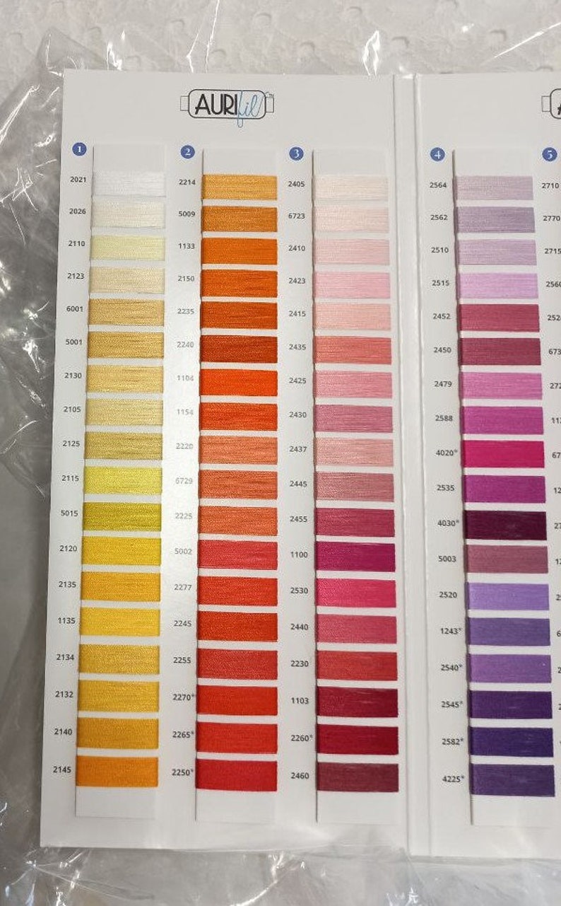 May include: An Aurifil thread color chart featuring a grid of numbered swatches in shades of yellow, orange, pink, red, and purple. The white card displays columns of thread samples arranged by color family, each labeled with a specific numerical code.