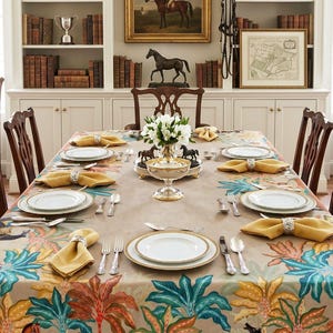May include: A rectangular dining table covered with a beige tablecloth featuring a colorful pattern of horses and palm trees. The table is set with white plates, gold-rimmed bowls, yellow napkins in silver rings, and polished silver cutlery.