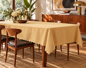 Puede incluir: Un mantel rectangular de color amarillo claro sólido, que presenta un diseño de borde festoneado decorativo. La tela cubre la parte superior de una mesa de comedor de madera y cae por los lados, proporcionando una superficie uniforme.