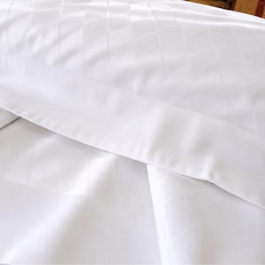 以下が含まれることがあります： 同系色のダイヤモンドジャカード模様が施された、鮮やかな白い枕カバーとシーツのセット。生地は滑らかで清潔感のある仕上がりで、端には幅広の無地の縁取りがあり、洗練された均一な白い質感が際立っています。