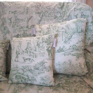 Puede incluir: Un coj&iacute;n cuadrado con base de tela blanca y un patr&oacute;n toile de jouy verde que representa escenas pastorales con figuras, &aacute;rboles y un perro. El coj&iacute;n tiene una peque&ntilde;a etiqueta rectangular en la costura lateral y una funda de tela a juego.