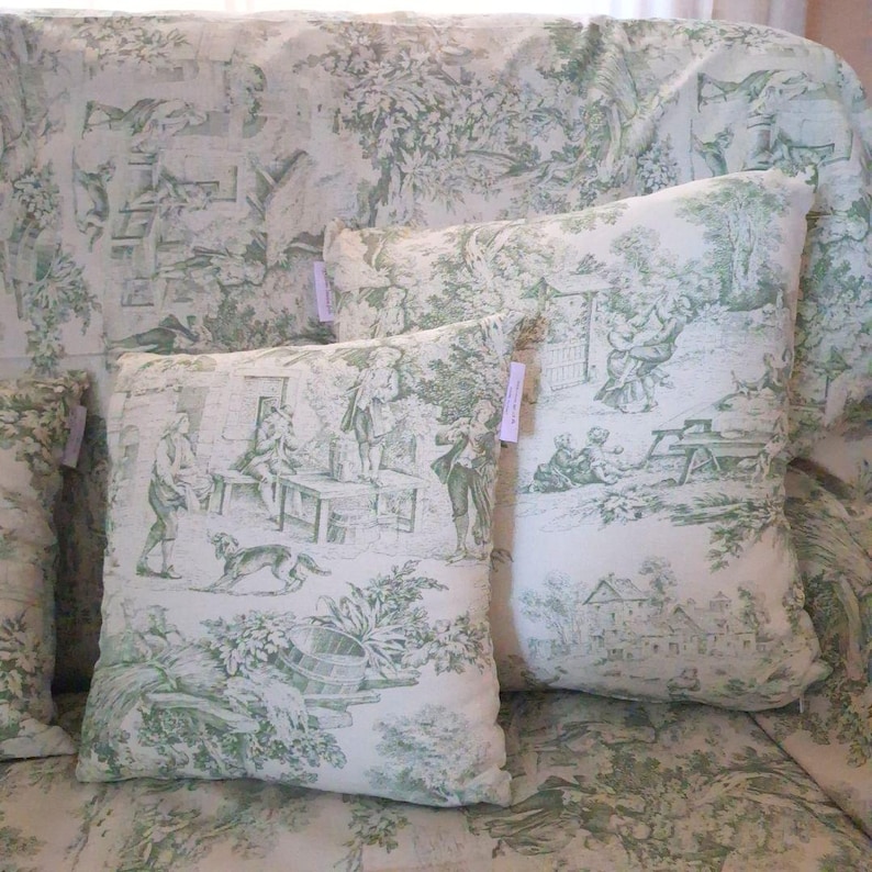 Puede incluir: Un coj&iacute;n cuadrado con base de tela blanca y un patr&oacute;n toile de jouy verde que representa escenas pastorales con figuras, &aacute;rboles y un perro. El coj&iacute;n tiene una peque&ntilde;a etiqueta rectangular en la costura lateral y una funda de tela a juego.