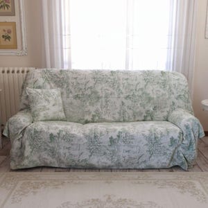 May include: A sofa covered in a white fabric featuring a repeating green toile pattern depicting pastoral scenes with trees and figures. The cover includes a matching square throw pillow placed on the left side of the seat, draping over the arms.