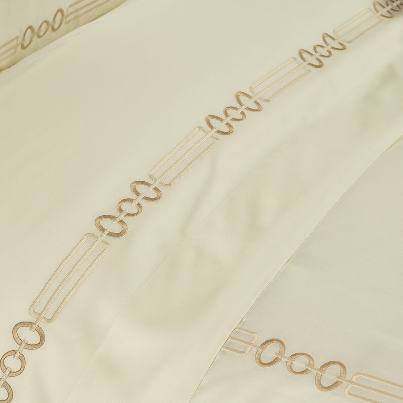 May include: A cream-colored fabric featuring an embroidered geometric pattern in a light tan thread. The design consists of repeating oval loops connected by elongated rectangular bars, creating a continuous linear motif across the surface.