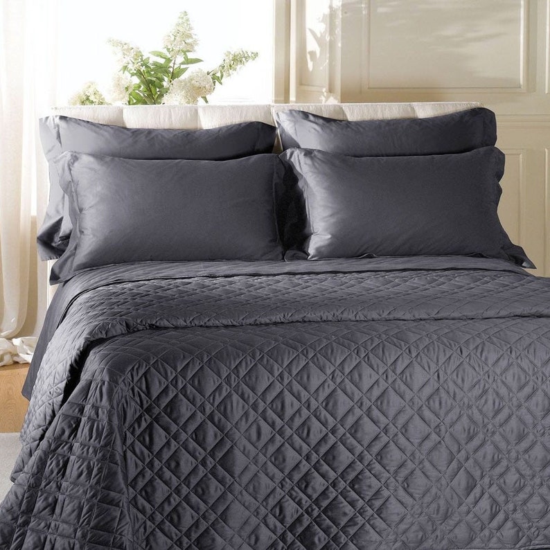 May include: A dark gray bedding set featuring a quilted coverlet with a diamond-stitched pattern and four matching rectangular pillowcases. The bedding is arranged neatly on a bed, showcasing a smooth, uniform dark gray color throughout the set.