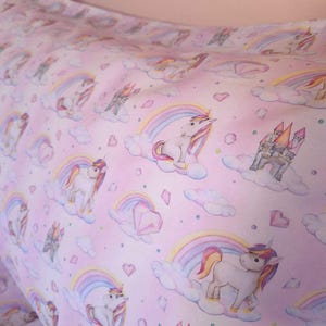 May include: Pink fabric with a repeating pattern of unicorns, rainbows, clouds, castles, and diamonds. The unicorns have colorful manes and tails. The rainbows have pastel colors. The overall design is whimsical and fantasy-themed.